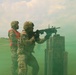 Two soldiers in U.S. Army camouflage uniforms are engaged in what appears to be a military training exercise on an open field. One soldier is firing or aiming a rifle (appears to be an M4/M16-style weapon) from behind a concrete barrier, while a second soldier wearing a high-visibility orange safety vest observes or supervises. Green smoke from a smoke grenade fills the surrounding area, indicating a tactical training scenario.