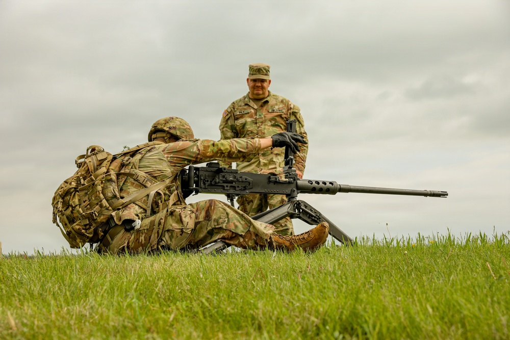 Two military personnel in MultiCam/OCP camouflage uniforms are engaged in what appears to be a weapons training exercise on an open grassy field. One soldier is in a prone position operating a large-caliber machine gun on a bipod, while a second soldier stands behind observing or instructing. The setting suggests a training range environment under overcast skies.