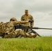 Two military personnel in MultiCam/OCP camouflage uniforms are engaged in what appears to be a weapons training exercise on an open grassy field. One soldier is in a prone position operating a large-caliber machine gun on a bipod, while a second soldier stands behind observing or instructing. The setting suggests a training range environment under overcast skies.