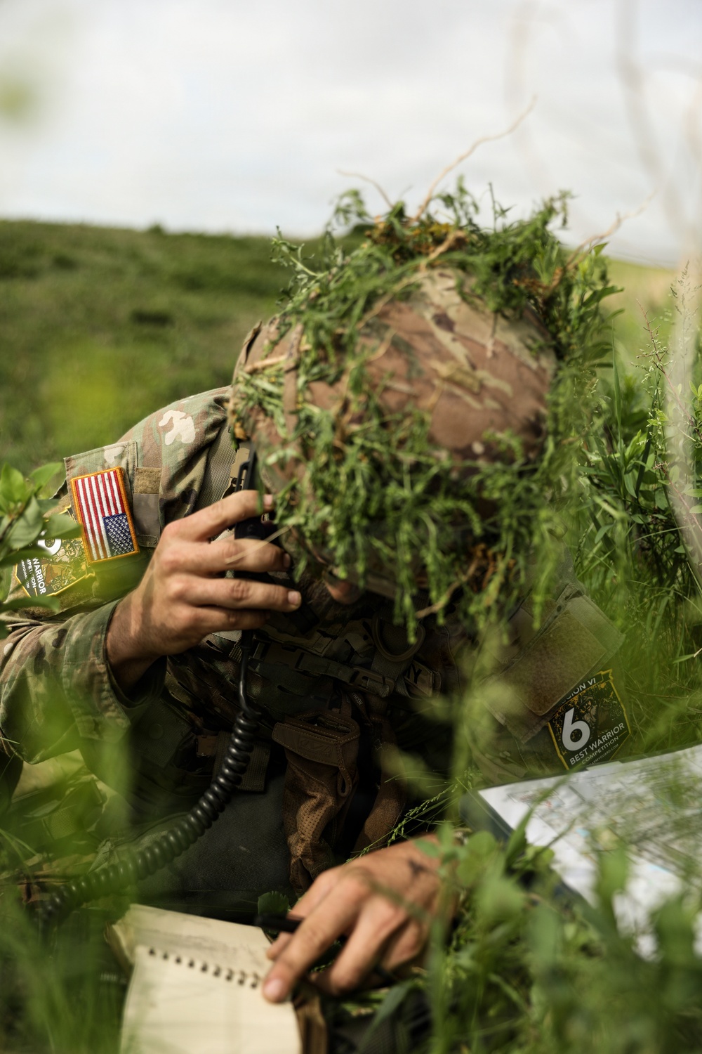A U.S. Army soldier in full camouflage gear, including a ghillie-style helmet cover with natural vegetation, is using a radio handset while referencing a map and notepad in an outdoor field setting. The soldier is participating in what appears to be the 'Best Warrior Competition' based on a visible patch on their gear. The face is completely obscured by the camouflage helmet cover and foliage.