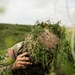 A U.S. Army soldier in full camouflage gear, including a ghillie-style helmet cover with natural vegetation, is using a radio handset while referencing a map and notepad in an outdoor field setting. The soldier is participating in what appears to be the 'Best Warrior Competition' based on a visible patch on their gear. The face is completely obscured by the camouflage helmet cover and foliage.