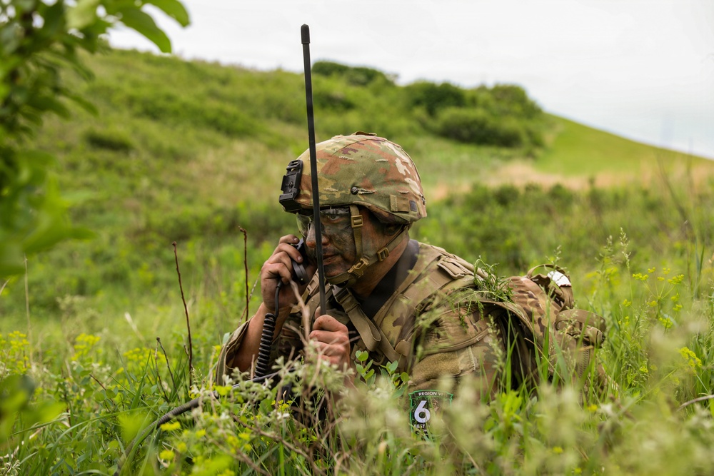 A soldier in full camouflage gear is lying prone in tall grass and vegetation, using a handheld military radio with an extended antenna. The soldier's face is painted with camouflage and they are wearing a tactical helmet with mounted equipment. A patch on the uniform reads 'Region 6' suggesting a unit or exercise designation.