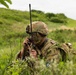 A soldier in full camouflage gear is lying prone in tall grass and vegetation, using a handheld military radio with an extended antenna. The soldier's face is painted with camouflage and they are wearing a tactical helmet with mounted equipment. A patch on the uniform reads 'Region 6' suggesting a unit or exercise designation.