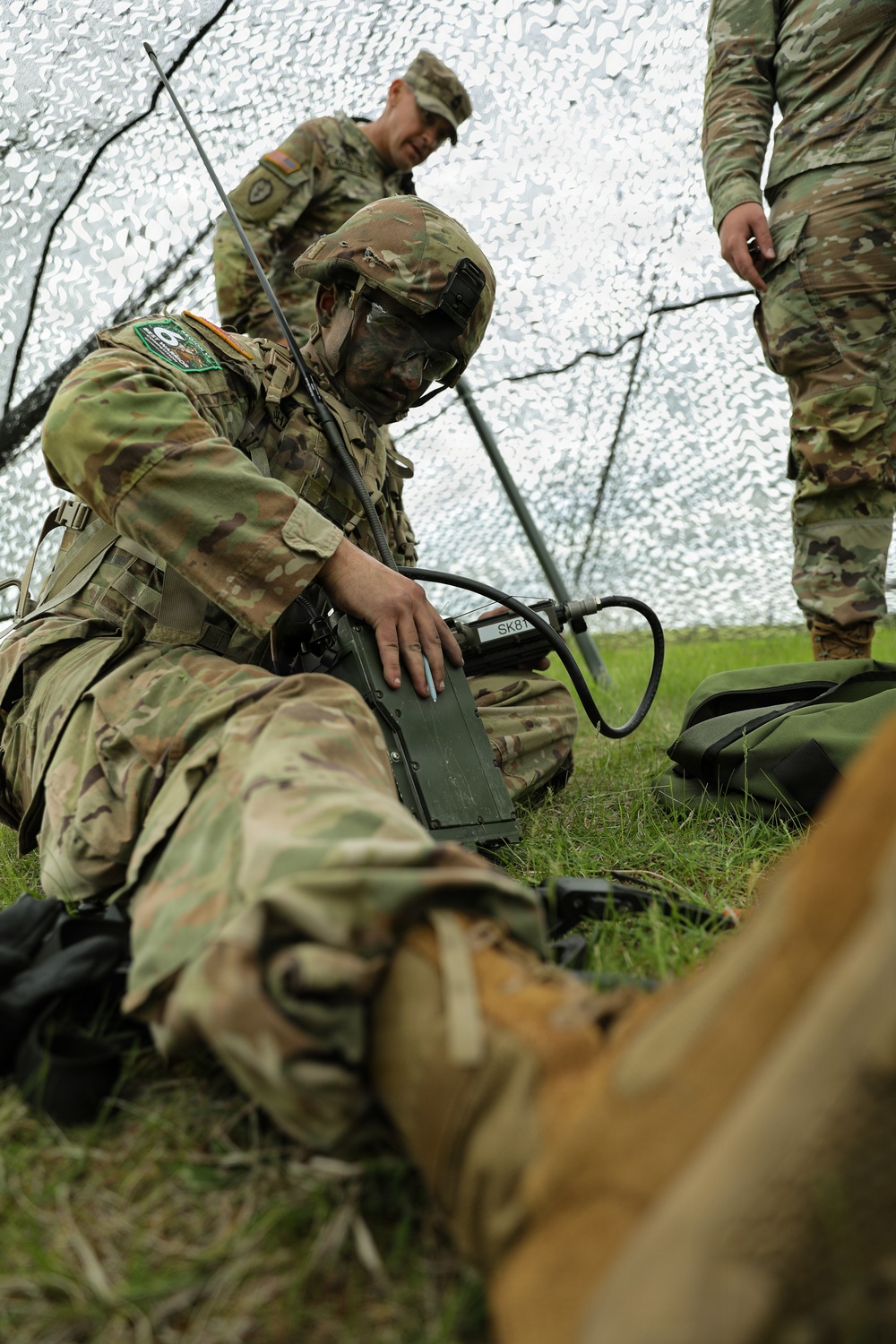 A soldier in full combat gear with face camouflage paint is operating a military radio/communications device on the ground beneath a camouflage net. Two additional soldiers in MultiCam (OCP) uniforms are present in the background. The setting appears to be a field training exercise or tactical operations scenario.