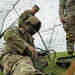 A soldier in full combat gear with face camouflage paint is operating a military radio/communications device on the ground beneath a camouflage net. Two additional soldiers in MultiCam (OCP) uniforms are present in the background. The setting appears to be a field training exercise or tactical operations scenario.