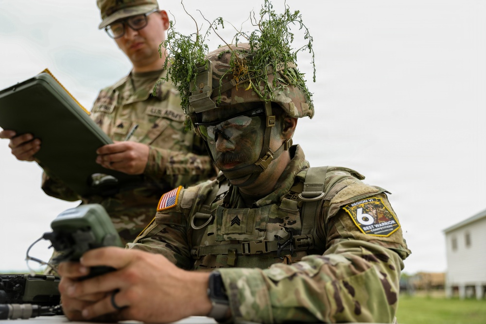 Two U.S. Army soldiers are engaged in a field training exercise or competition. The foreground soldier, wearing camouflage face paint and natural foliage on his helmet, is operating a military handheld device. A second soldier in the background is reviewing a tablet or clipboard, likely serving as an evaluator or observer.