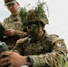 Two U.S. Army soldiers are engaged in a field training exercise or competition. The foreground soldier, wearing camouflage face paint and natural foliage on his helmet, is operating a military handheld device. A second soldier in the background is reviewing a tablet or clipboard, likely serving as an evaluator or observer.