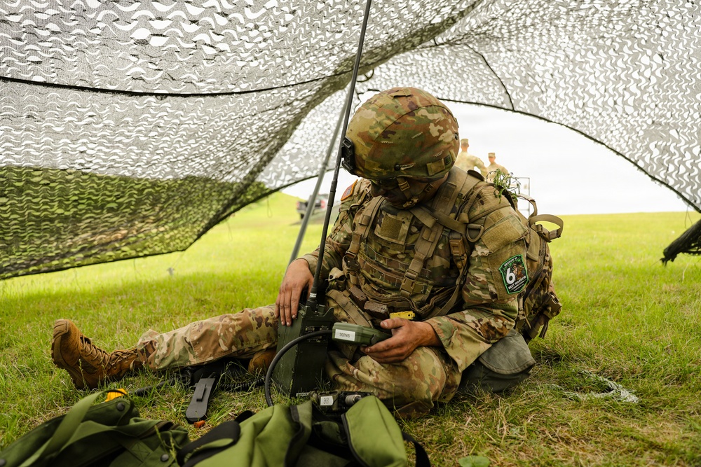 A soldier in full combat gear is seated on grass beneath a camouflage netting, operating what appears to be a military radio communications device with an antenna. The soldier is wearing Operational Camouflage Pattern (OCP) uniform, a tactical helmet, body armor, and a unit patch visible on the left shoulder. Additional personnel are visible in the background in an open field setting.