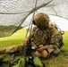 A soldier in full combat gear is seated on grass beneath a camouflage netting, operating what appears to be a military radio communications device with an antenna. The soldier is wearing Operational Camouflage Pattern (OCP) uniform, a tactical helmet, body armor, and a unit patch visible on the left shoulder. Additional personnel are visible in the background in an open field setting.