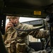 A U.S. Army Sergeant is seated inside a military vehicle, likely a HMMWV (Humvee), using a coiled-cord radio handset to communicate. The soldier is wearing Operational Camouflage Pattern (OCP) uniform with full tactical gear including a plate carrier and MOLLE pouches. A yellow caution label warning about high-intensity noise and hearing protection is visible on the vehicle interior.