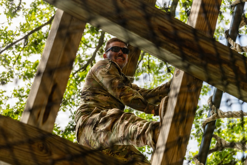 A soldier in camouflage uniform is climbing a wooden obstacle course structure, photographed from a low angle looking upward. The individual appears to be smiling and wearing dark sunglasses while navigating the wooden beams and rope elements. The setting appears to be an outdoor military training facility surrounded by trees.
