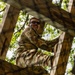 A soldier in camouflage uniform is climbing a wooden obstacle course structure, photographed from a low angle looking upward. The individual appears to be smiling and wearing dark sunglasses while navigating the wooden beams and rope elements. The setting appears to be an outdoor military training facility surrounded by trees.