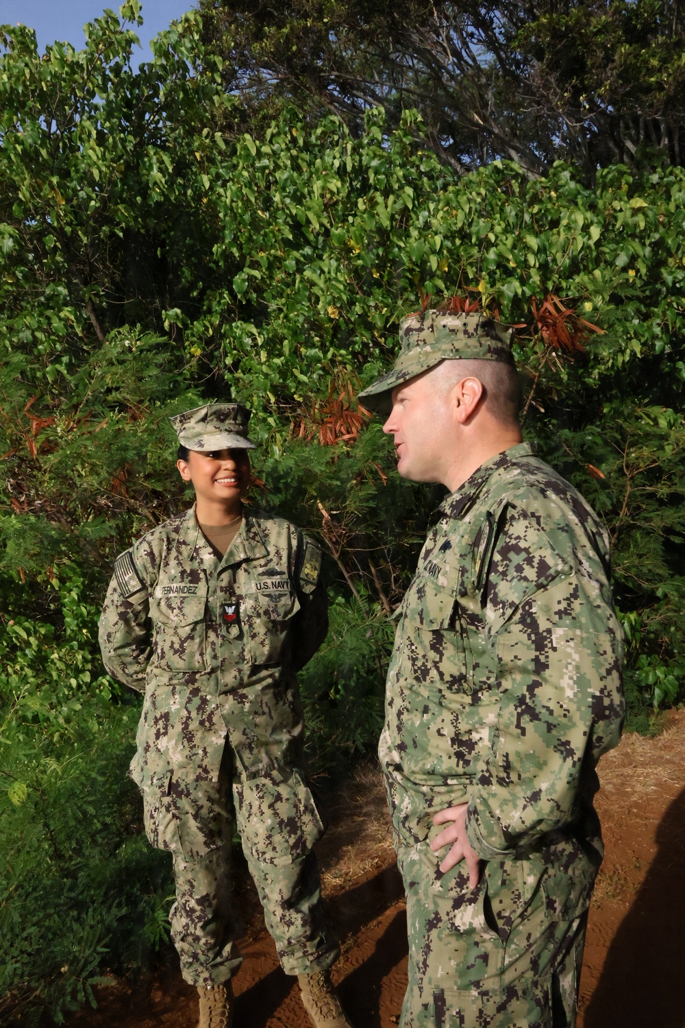 Two U.S. Navy personnel in NWU (Navy Working Uniform) Type III camouflage are engaged in conversation outdoors. The sailor on the left, identified by name tape as 'Fernandez,' is smiling and appears to be speaking with a higher-ranking male sailor on the right. The setting appears to be a tropical or subtropical outdoor environment with dense green vegetation.