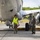 Two military personnel are conducting a pre-flight or post-flight inspection of a large military aircraft on a tarmac. One individual wearing a high-visibility yellow vest and hearing protection is inspecting the underside of the aircraft fuselage, while a second person in an olive green flight suit and helmet stands nearby observing. The setting appears to be a tropical or subtropical airbase based on the lush green vegetation visible in the background.