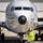 A U.S. Navy aircraft handler in a high-visibility safety vest and flight deck helmet is marshaling a military patrol aircraft (numbered 325) on a tarmac. The individual is facing the aircraft with both arms raised, giving directional hand signals to guide the aircraft. The aircraft appears to be a Boeing P-8 Poseidon maritime patrol aircraft based on its distinctive nose radome and fuselage design.