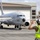 A military aircraft handler in a high-visibility vest and flight deck helmet is marshaling a U.S. Navy P-8 Poseidon maritime patrol aircraft (bureau number 325) on a tarmac. The handler's raised hands indicate an active marshaling signal being communicated to the flight crew. The scene appears to take place at a military air station with large hangars visible in the background.