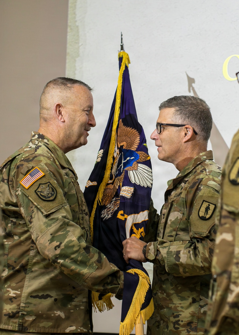 Two U.S. Army soldiers in OCP (Operational Camouflage Pattern) uniforms are participating in what appears to be a change of command or guidon transfer ceremony. They are jointly holding a dark blue military guidon/flag adorned with an eagle emblem and gold fringe. The soldier on the left is smiling, suggesting a formal but positive ceremonial exchange.