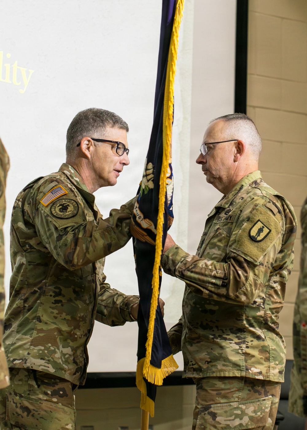 Two U.S. Army soldiers in OCP (Operational Camouflage Pattern) uniforms are participating in a military change of command or activation ceremony, passing a unit guidon/colors between them. The ceremony appears to be taking place indoors, likely in a military facility or armory. The passing of the flag is a formal military tradition symbolizing transfer of authority or responsibility.