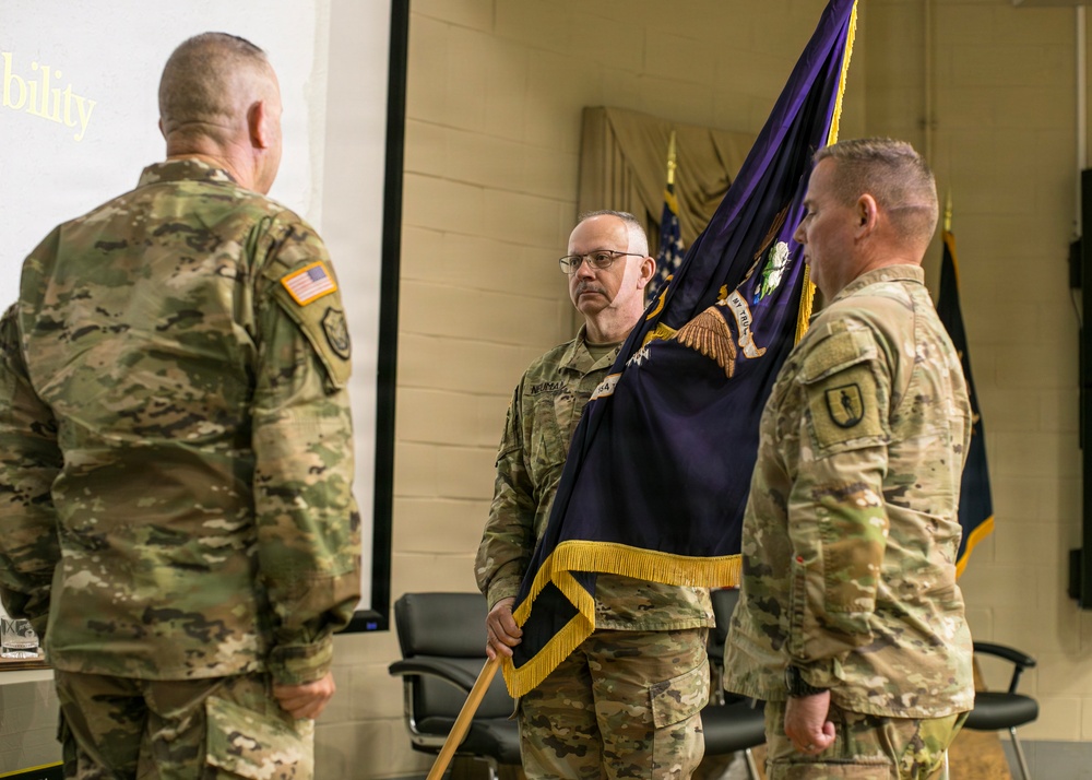 Three U.S. Army soldiers in OCP (Operational Camouflage Pattern) uniforms are participating in what appears to be a military change of command or activation ceremony. The central figure is holding a purple and gold ceremonial guidon or unit colors, which is a hallmark of formal military ceremonies. The setting is an indoor military facility with a projection screen and additional flags visible in the background.