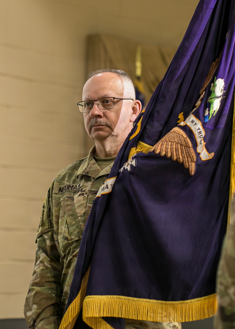 A U.S. Army soldier in OCP (Operational Camouflage Pattern) uniform is holding a purple and gold military guidon or unit flag during what appears to be a formal military ceremony. The soldier, identified by the name tape 'NEUMAN' and a partially visible unit designation '154T' or similar, stands at attention in an indoor venue. The flag features an eagle emblem and gold fringe, consistent with a U.S. Army unit colors flag.