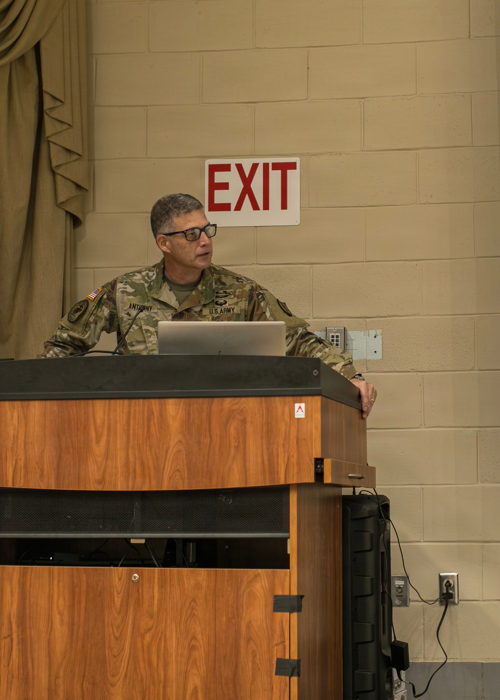 A U.S. Army soldier identified by name tape as 'ANTHONY' stands at a wooden podium delivering a briefing or presentation. He is wearing OCP (Operational Camouflage Pattern) uniform with U.S. Army branch tape, an American flag patch, and additional unit patches. A laptop is open on the podium in front of him, and an EXIT sign is visible on the cinder block wall behind him.