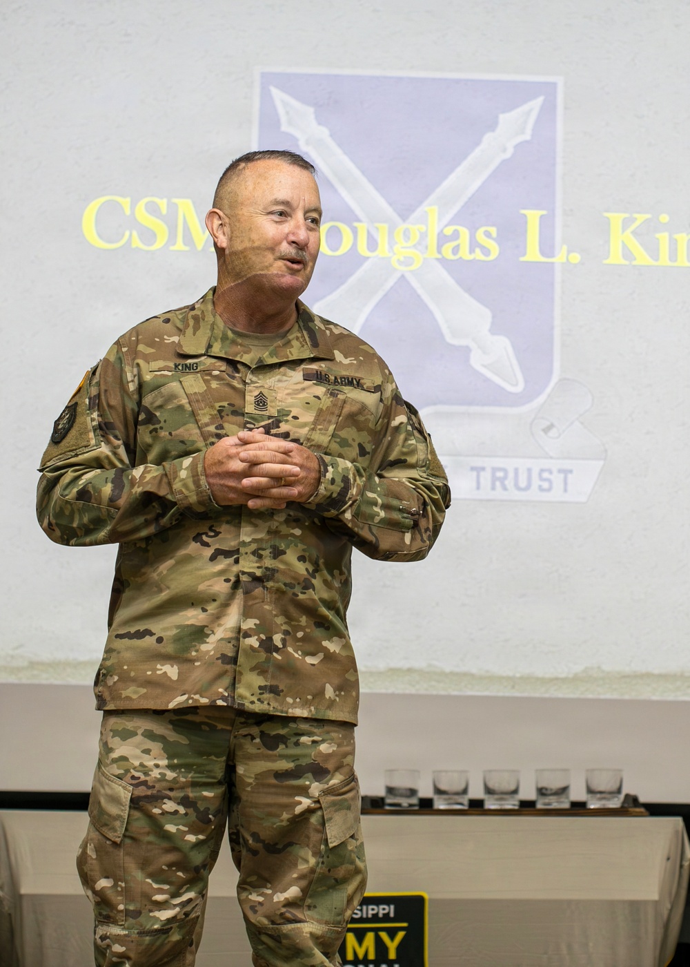 A U.S. Army soldier in OCP (Operational Camouflage Pattern) uniform stands before a projected presentation slide. The projection behind him displays 'CSM Douglas L. Ki[ng]' along with a military unit crest and the word 'TRUST.' He appears to be addressing an audience at a formal military event or ceremony.