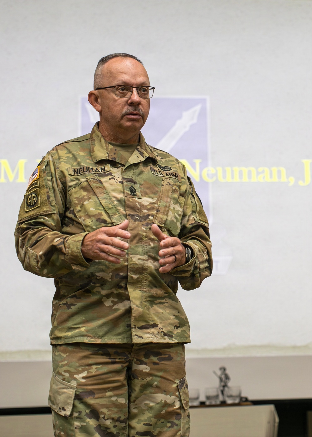 A U.S. Army senior non-commissioned officer named Neuman is delivering a presentation or briefing in front of a projected screen. He is wearing the Army Combat Uniform (ACU) in MultiCam/OCP pattern with visible rank insignia and unit patches. The projected slide behind him displays his name and what appears to be a unit insignia.