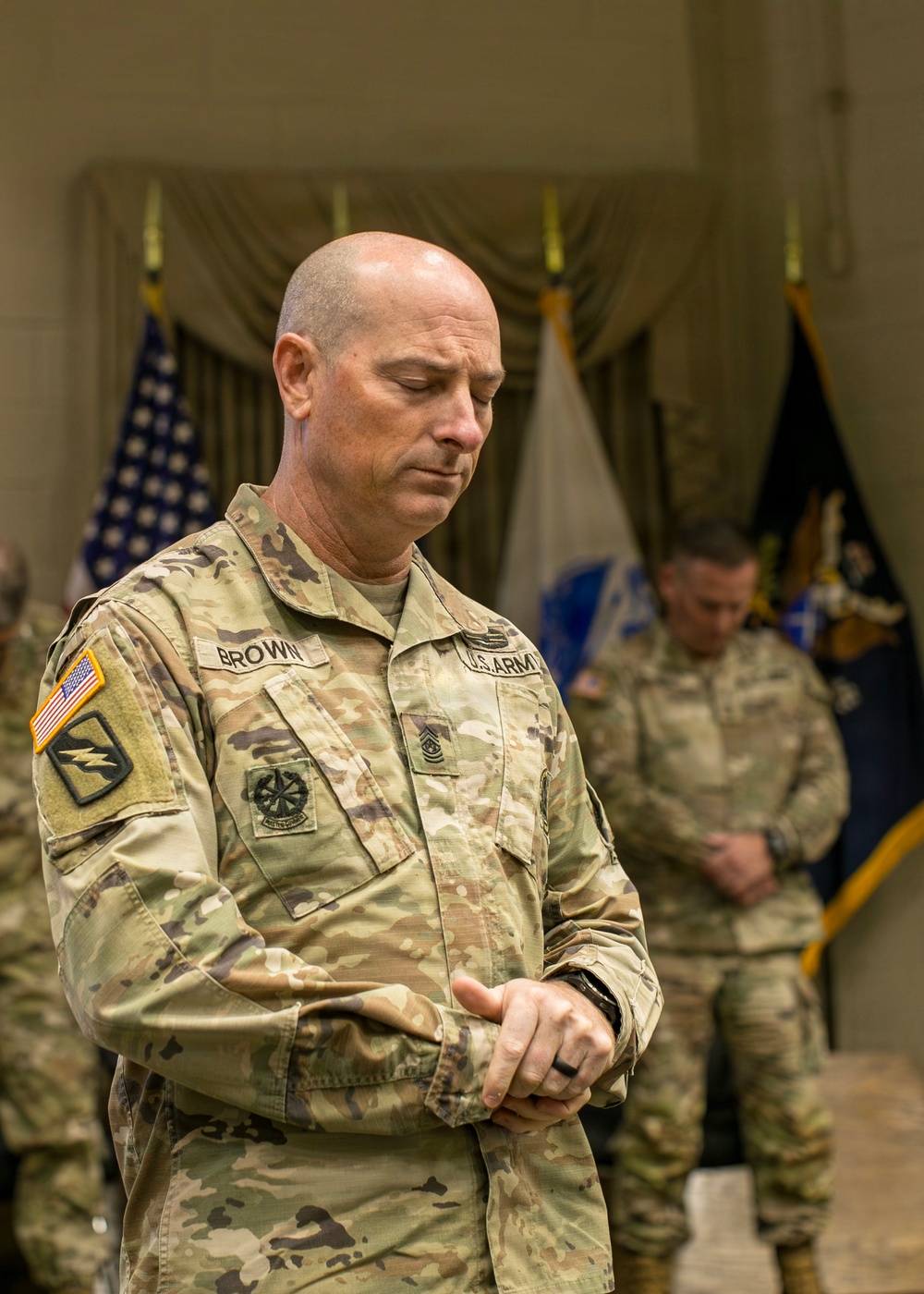 A U.S. Army soldier with the name tape 'BROWN' and senior enlisted rank insignia stands with head bowed and eyes closed, appearing to be in prayer or a moment of reflection during a formal military ceremony. Multiple flags are visible in the background, along with at least one other soldier also in a bowed posture. The setting appears to be an indoor ceremonial hall.