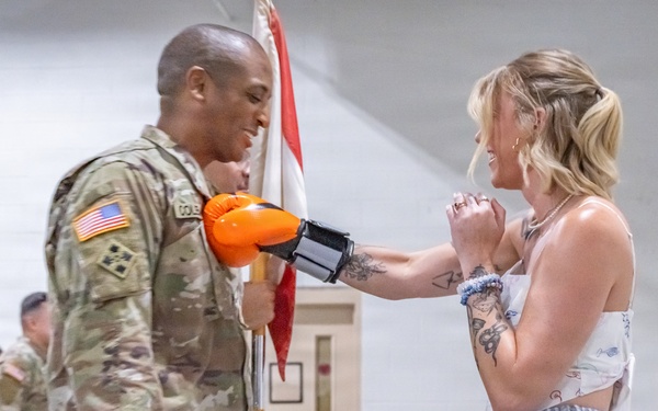A U.S. Army soldier in OCP (Operational Camouflage Pattern) uniform is interacting with a civilian woman who is wearing an orange boxing glove on one hand. The soldier appears to be smiling and leaning in toward the woman, who is holding his gloved hand with both of hers. The scene takes place inside what appears to be a military hangar or gymnasium, with other uniformed soldiers visible in the background.