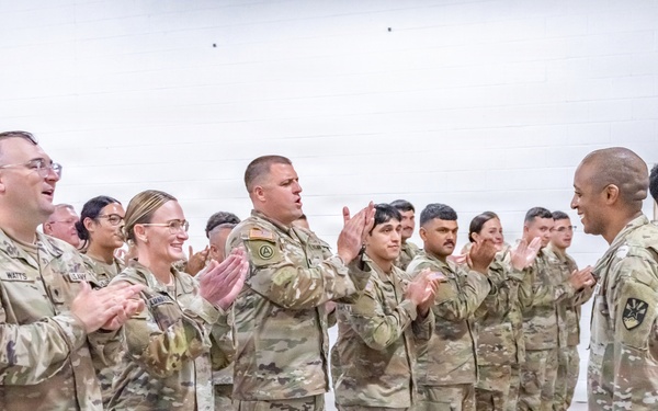 A group of U.S. Army soldiers in OCP (Operational Camouflage Pattern) uniforms are gathered in an indoor facility, applauding during what appears to be a military ceremony or formation. Two soldiers stand facing the group on the right side, one holding a unit guidon (flag on a staff). The setting appears to be a drill hall or armory with a white concrete block wall background.