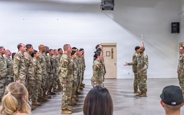 A military formation ceremony is taking place inside a large indoor facility with white cinder block walls. A group of soldiers in Army Combat Uniforms (ACU/OCP pattern) are standing in formation while two or three senior personnel address them. Civilian observers, likely family members, are seated in folding chairs in the foreground watching the proceedings.