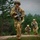 Three soldiers in full combat gear are conducting a patrol or field exercise in a forested, hilly terrain under overcast skies. The lead soldier is moving forward carrying an assault rifle with an underbarrel grenade launcher attachment, while two soldiers follow behind in staggered formation. All personnel are wearing MultiCam (OCP) camouflage uniforms consistent with U.S. Army equipment standards.