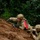 Two soldiers in full combat gear are positioned behind a dirt embankment in a wooded training environment. The lead soldier is pointing forward and appears to be issuing directions or identifying a target, while the second soldier holds a rifle in a ready position. Both wear MultiCam (OCP) uniforms with tactical helmets and carry rifles, suggesting a military field training exercise.