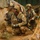 Three U.S. Army soldiers in full combat gear are moving through a narrow trench or defile during what appears to be a field training exercise. The soldiers are wearing MultiCam (OCP) uniforms, tactical helmets, and face camouflage paint, and are armed with M4-style carbines. The lead soldier is holding what appears to be a smoke grenade or similar device.