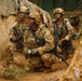 Three U.S. Army soldiers in full combat gear are moving through a narrow trench or defile during what appears to be a field training exercise. The soldiers are wearing MultiCam (OCP) uniforms, tactical helmets, and face camouflage paint, and are armed with M4-style carbines. The lead soldier is holding what appears to be a smoke grenade or similar device.
