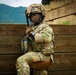 A U.S. Army soldier in full combat gear is positioned against a wooden barrier, holding a rifle in a tactical stance. The soldier is wearing Operational Camouflage Pattern (OCP) uniform with a camouflaged helmet, protective eyewear, and a communication headset. The setting appears to be a military training exercise in a forested, mountainous environment.