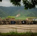 A large group of military personnel pose for a unit photo between two armored vehicles in a mountainous, forested setting. Two unit flags are visible being held in the center of the formation, with the numbers '23' and '4' visible on one flag. The setting appears to be a training area or field exercise location.