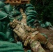 A soldier in full multicam camouflage uniform and tactical vest is lying prone on the ground beside a fortified sandbag position, with one arm extended upward toward the sandbag wall. The setting appears to be a wooded or vegetated area with green woven sandbags stacked as a defensive barrier. A wooden pallet is visible atop the sandbag structure.