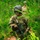 Two soldiers in full combat gear move through a dense pine forest during what appears to be a field training exercise. The lead soldier has green and black camouflage face paint applied and carries an M4-style carbine rifle. Both soldiers wear OCP (Operational Camouflage Pattern) uniforms with tactical vests and helmets.