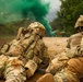 Two soldiers in full combat gear are lying prone on a dirt ground, facing toward a green smoke signal in the distance. The scene appears to be a military training exercise or tactical operation, with green smoke grenades deployed approximately 30-40 meters ahead. Dense vegetation lines the background behind the smoke.