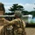 A soldier in full camouflage gear is firing a shoulder-launched rocket or recoilless weapon system from a fortified wooden position. Backblast smoke is clearly visible emanating from the rear of the weapon tube. The setting appears to be a military training range with trees and a gravel surface visible in the background.
