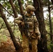 A fully equipped soldier in multicam/OCP camouflage uniform stands among trees in a forested environment. The individual is carrying an assault rifle and wearing full tactical gear including a helmet with mounted accessories, plate carrier, and communications equipment. The setting appears to be a training exercise or patrol in a woodland area.