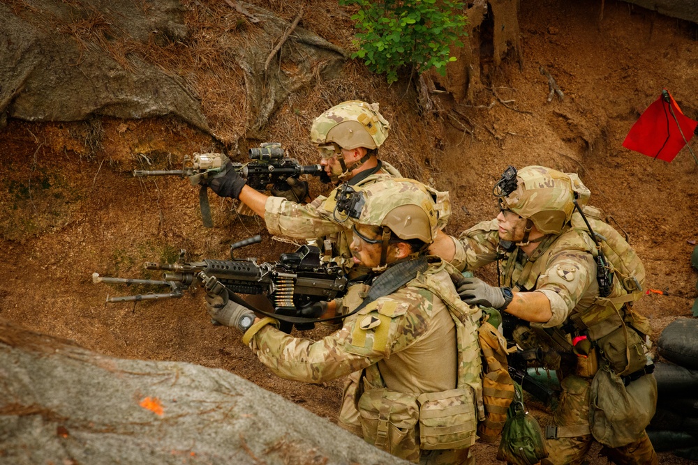 Three soldiers in full combat gear are positioned in a rocky, earthen terrain, aiming weapons in a coordinated tactical formation. They are wearing matching multicam/OCP camouflage uniforms with helmets, tactical vests, and gloves. A red marker flag is visible in the upper right corner, suggesting a training exercise or marked position.