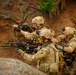Three soldiers in full combat gear are positioned in a rocky, earthen terrain, aiming weapons in a coordinated tactical formation. They are wearing matching multicam/OCP camouflage uniforms with helmets, tactical vests, and gloves. A red marker flag is visible in the upper right corner, suggesting a training exercise or marked position.