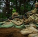 Three soldiers in MultiCam/OCP camouflage uniforms are positioned behind a sandbag fortification in a wooded area. The primary weapon visible is an M240 machine gun with a belt of ammunition, manned by a prone soldier, while a second soldier assists and a third observes from a kneeling position. The scene appears to be a defensive fighting position, likely during a training exercise.