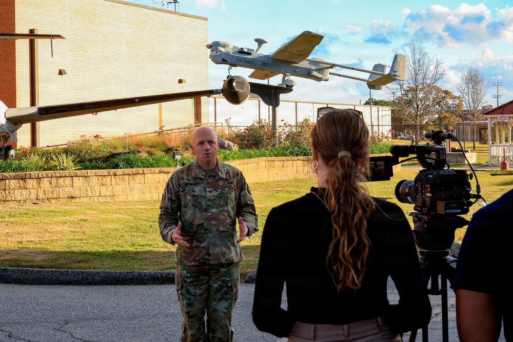 A U.S. Army soldier in camouflage uniform is being interviewed by a female journalist with a professional video camera on a tripod, likely at a military installation. In the background, a static display of what appears to be an RQ-7 Shadow unmanned aerial vehicle (UAV) numbered '303' is mounted on a pole as a monument or exhibit. The setting appears to be an outdoor area of a military base with landscaped grounds and brick buildings.