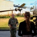A U.S. Army soldier in camouflage uniform is being interviewed by a female journalist with a professional video camera on a tripod, likely at a military installation. In the background, a static display of what appears to be an RQ-7 Shadow unmanned aerial vehicle (UAV) numbered '303' is mounted on a pole as a monument or exhibit. The setting appears to be an outdoor area of a military base with landscaped grounds and brick buildings.