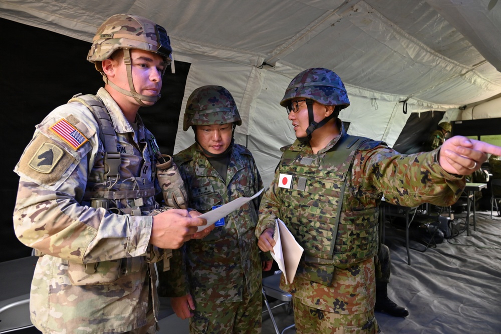 Three military personnel — one U.S. Army soldier and two Japan Ground Self-Defense Force (JGSDF) soldiers — are gathered under a military tent, reviewing documents together. The JGSDF soldier on the right is pointing in a direction while holding papers, suggesting a briefing or coordination activity. This appears to be a joint military exercise or operation between U.S. and Japanese forces.