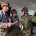 Three military personnel — one U.S. Army soldier and two Japan Ground Self-Defense Force (JGSDF) soldiers — are gathered under a military tent, reviewing documents together. The JGSDF soldier on the right is pointing in a direction while holding papers, suggesting a briefing or coordination activity. This appears to be a joint military exercise or operation between U.S. and Japanese forces.