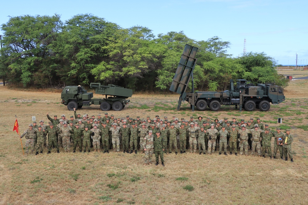 A large group of military personnel from what appears to be two different nations pose together in a field for a group photograph. Behind them are two military rocket/missile launcher systems mounted on heavy tactical trucks. The setting appears to be an open training area with trees in the background, likely during a joint military exercise.